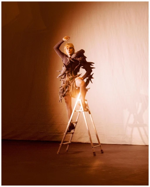 Fashion photo of a blonde adult female model in an avant-garde dress, climbing a ladder in a brightly lit photo studio setting.
