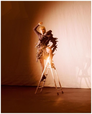 Fashion photo of a blonde adult female model in an avant-garde dress, climbing a ladder in a brightly lit photo studio setting.