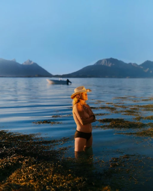Adult woman standing in water, wearing a hat and bottoms, mountains and a boat are visible in the distance under a clear sky.