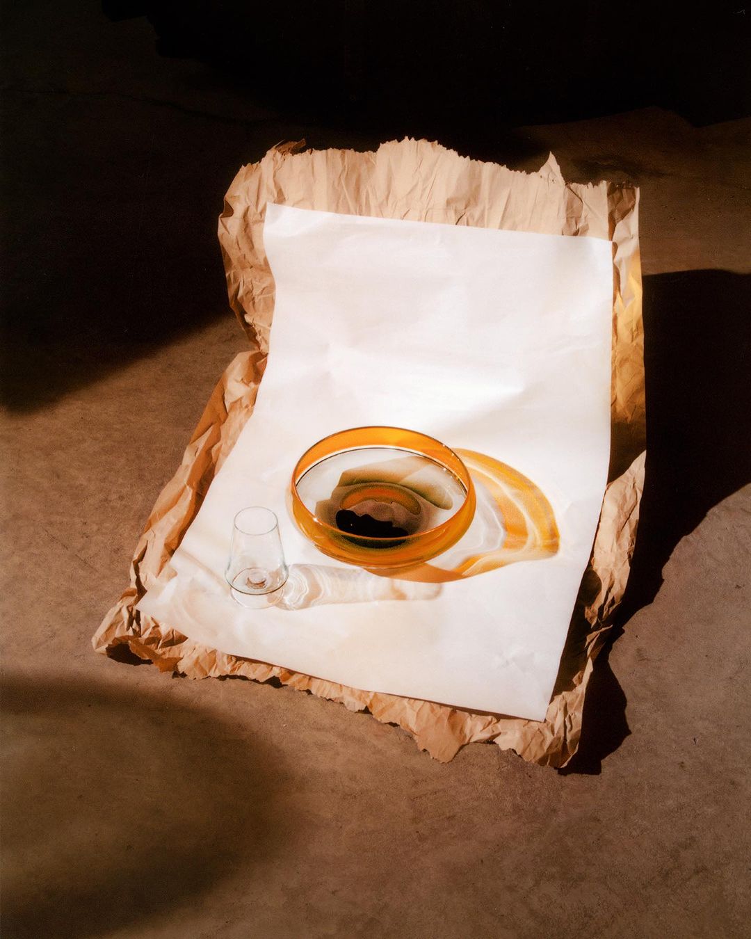Still life with empty drinking glass, clear orange-rimmed bowl with contents, and white and brown paper on a brown surface.