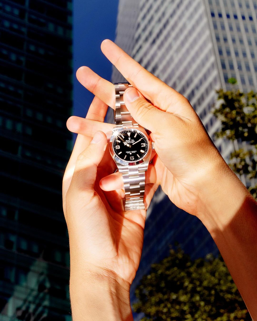 Close-up of a silver wristwatch held in two hands, presented against an outdoor backdrop of modern high-rise buildings and green trees.