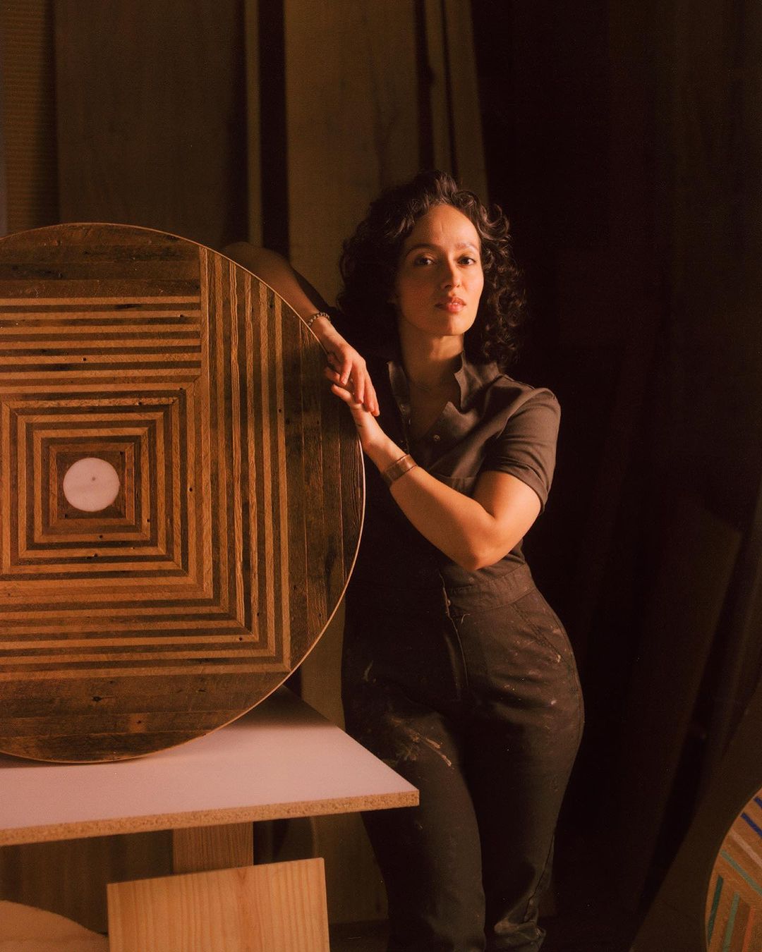 Adult female artist stands in her woodshop, leaning against a table she made with an eye-catching geometric wood pattern.
