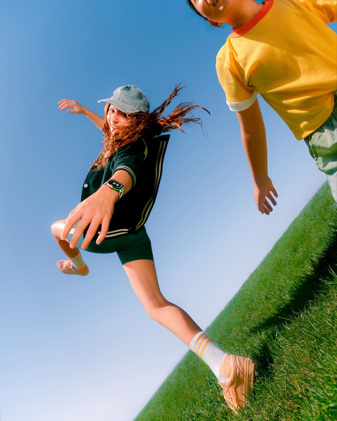 A young woman jumps with arms outstretched toward another person against a sky and grass background in a dynamic, angled shot.