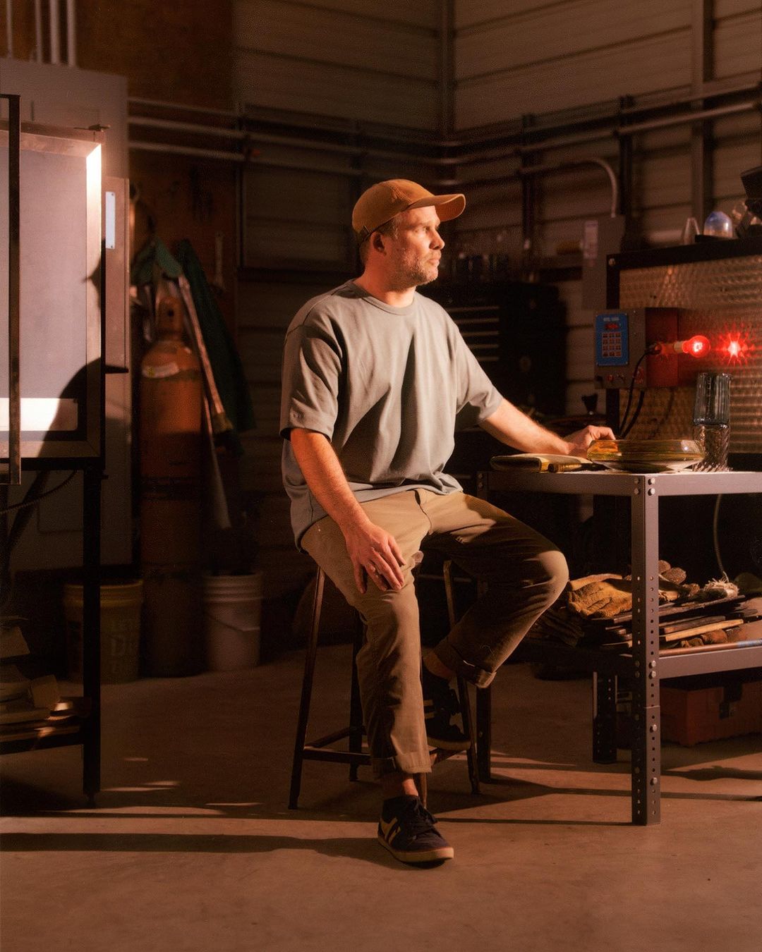 Man sitting in a workshop, wearing a t-shirt and cap, looking to the side with machinery, tools, and lighting in the background.