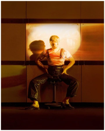 Blonde adult sits on a stool indoors. The person wears shiny pants, black shoes and a red striped T-shirt in a studio setting.