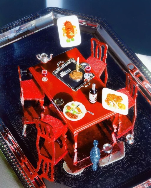 Miniature dining scene featuring a red table and chairs set on a silver tray with tiny plates of food and a wine bottle.