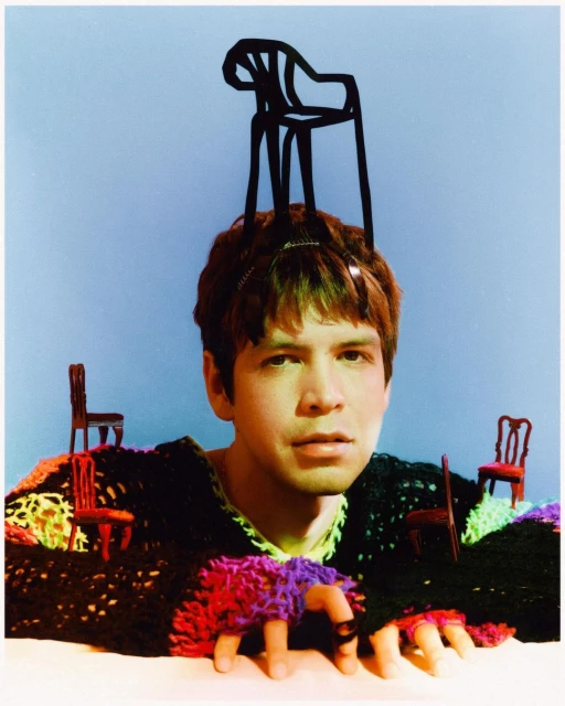 Portrait of a fair-skinned person with dark hair, wearing a multi-colored sweater and a chair on their head, against a blue background.
