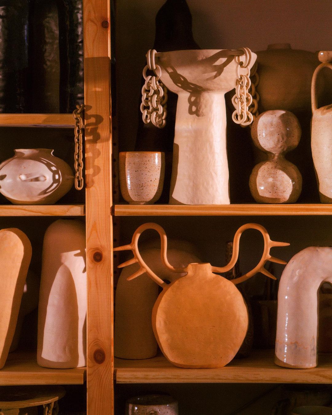 Various handcrafted ceramic pottery pieces on a wooden shelf, showcasing different shapes, textures, and beige tones, with artistic design.