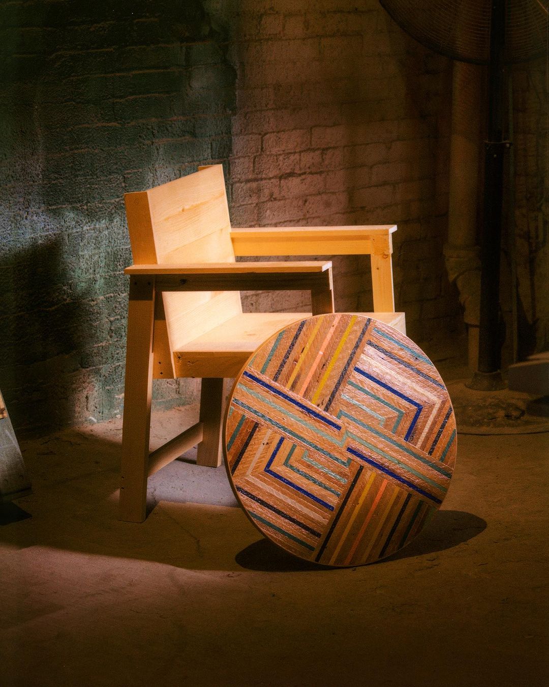 An unfinished wooden chair is positioned near a colorful patterned round table in a dimly lit space, featuring exposed brick and concrete.