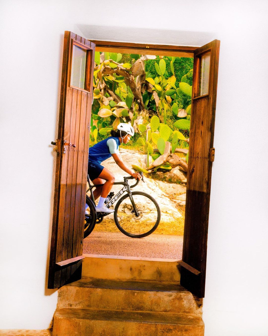 Cyclist in helmet and athletic clothing rides bike through open doorway into a sunlit, cactus-filled landscape with a white wall.