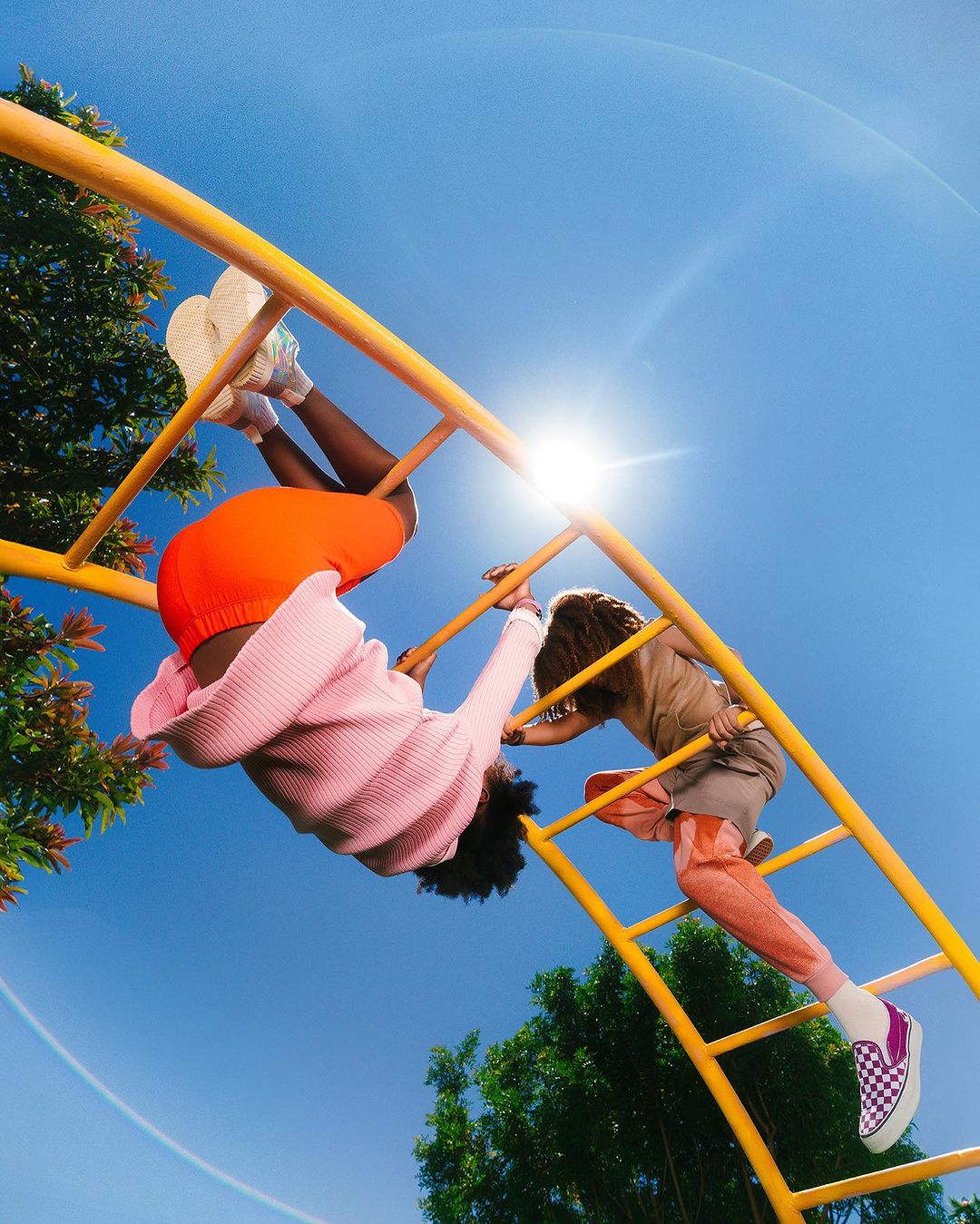 Two children playing on a yellow metal jungle gym under a sunny blue sky with green trees, one child hanging upside down.