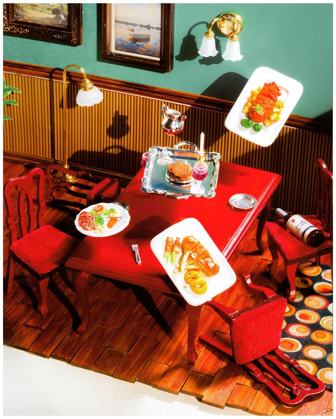 Overhead shot of a miniature dollhouse dining room featuring a red table, chairs, dishes of food, and decorative paintings and sconces.