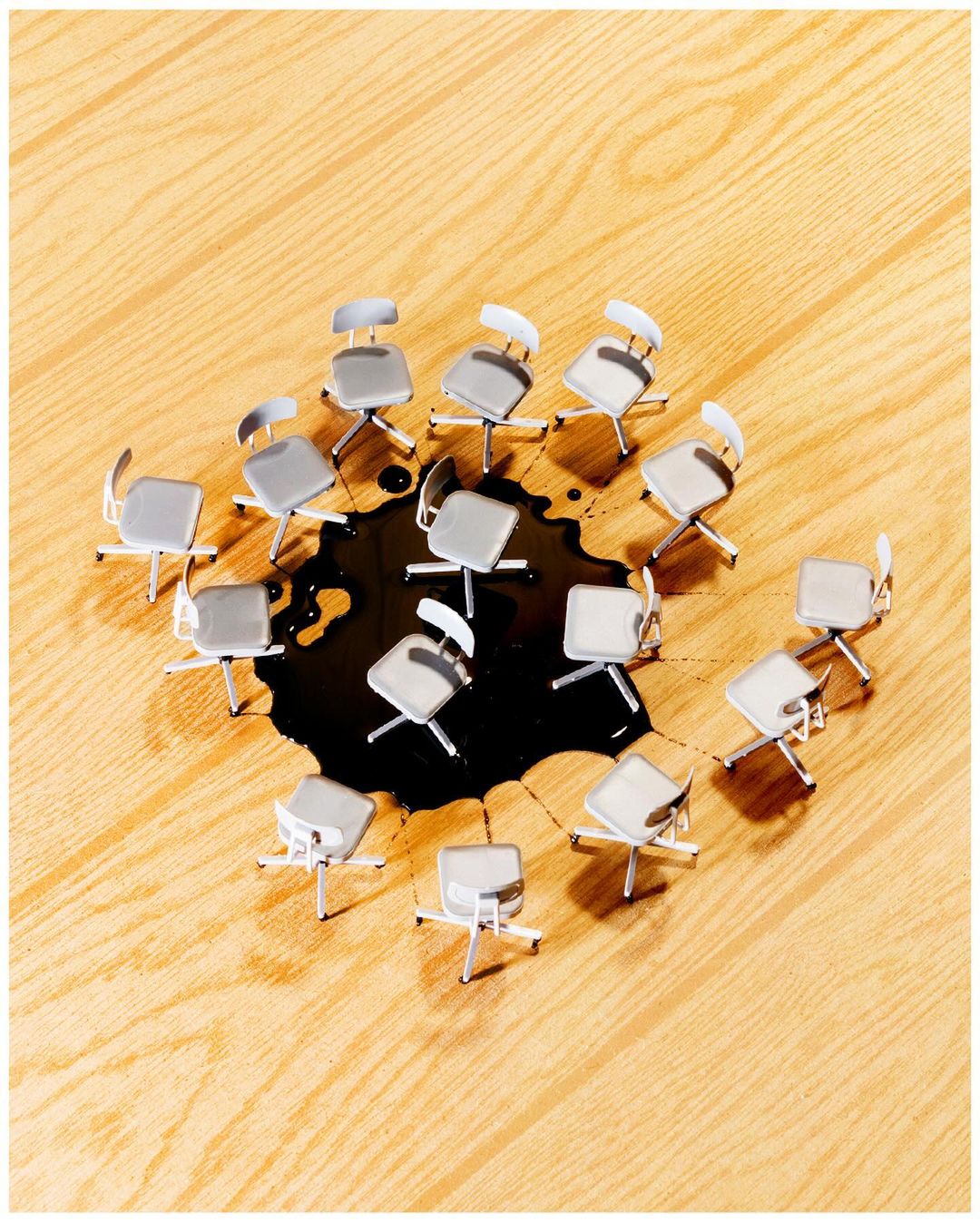 Overhead shot shows office chairs arranged in a circle around a dark liquid spill on a light wood floor, creating an interesting visual.