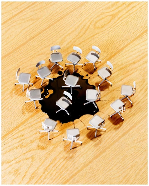Overhead shot shows office chairs arranged in a circle around a dark liquid spill on a light wood floor, creating an interesting visual.