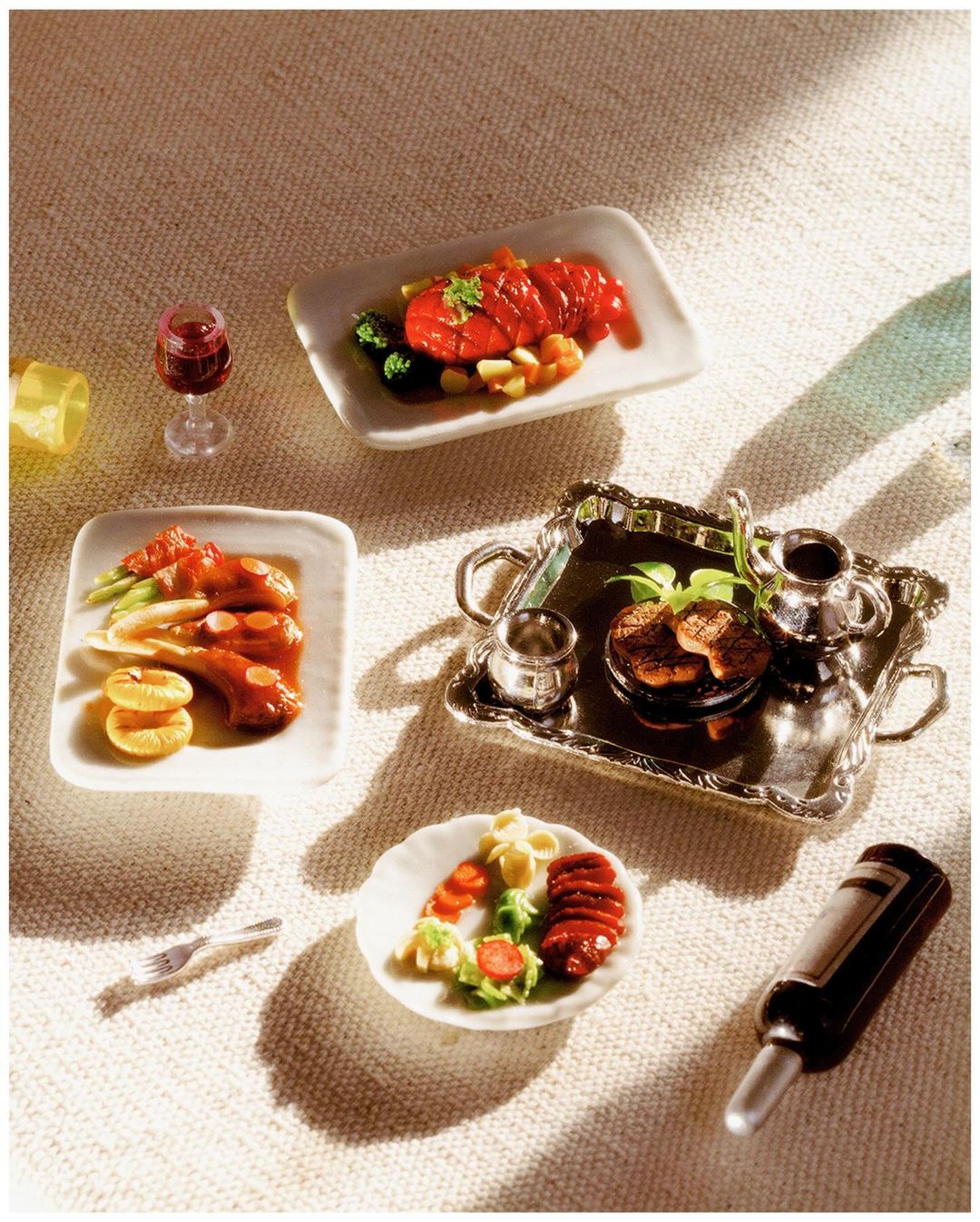 Overhead shot of miniature food dishes on a textured white surface featuring small portions of meat, salad, and drinks.