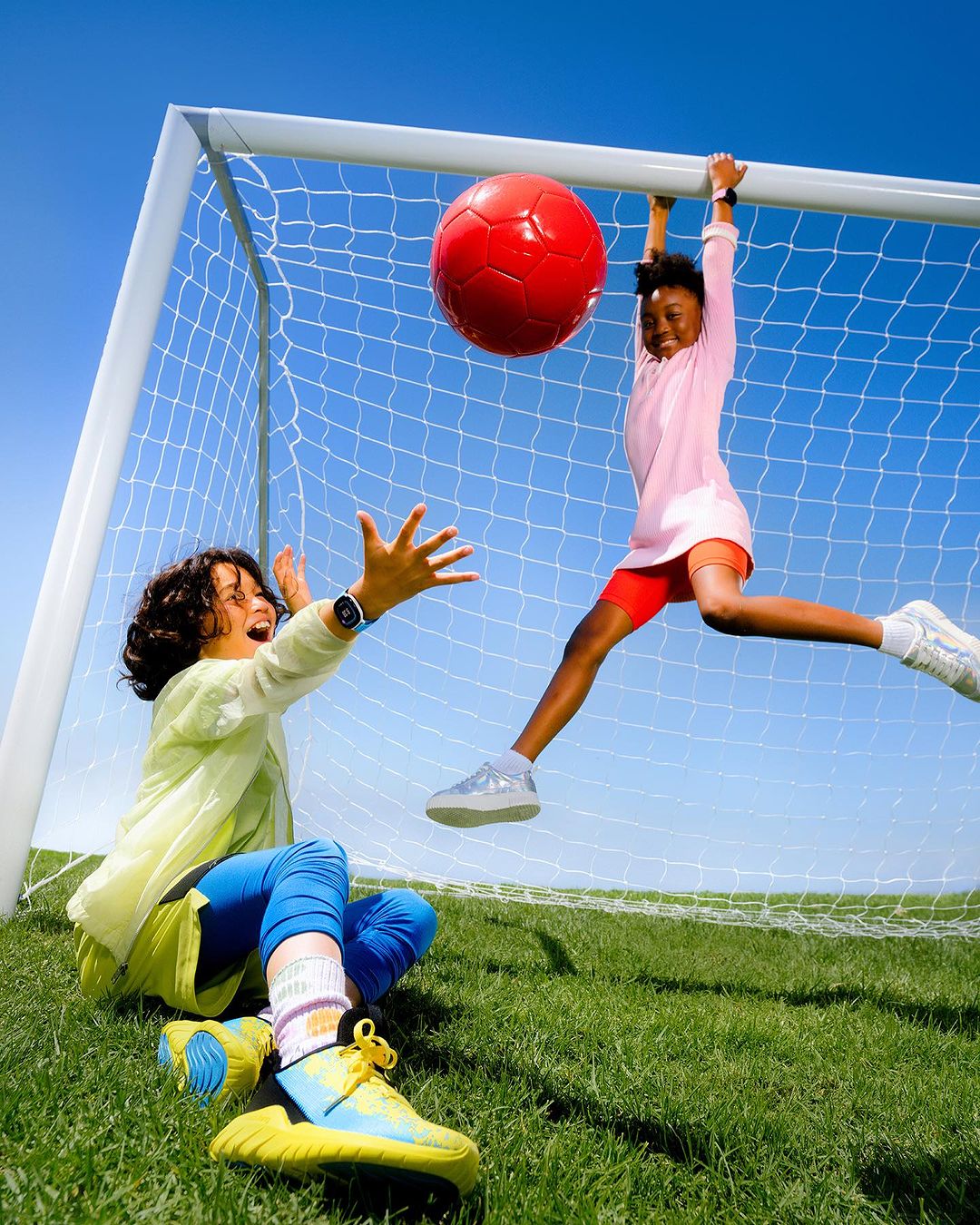 Two kids playing with a large red ball near a soccer goal on a sunny day with a clear blue sky and green grass.