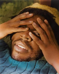 Close-up portrait of a Black man with a nose ring and blue textured sweater, holding hands over his face, photographed by Tonje Thilesen.