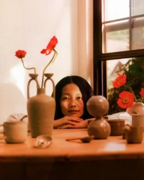 Woman resting her chin on her hands behind a wooden table filled with ceramic vases and red flowers by Tonje Thilesen
