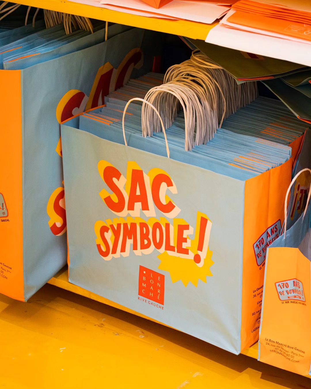 Stacks of shopping bags with orange and light blue color scheme are stacked on yellow shelving inside a store.