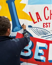 Illustrator Toque Freres painting a colourful mural with blue and red lettering in Paris