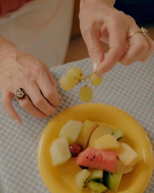Close-up of senior woman's hands holding grapes, with a plate of fruit on a blue gingham tablecloth in a warm, inviting setting.