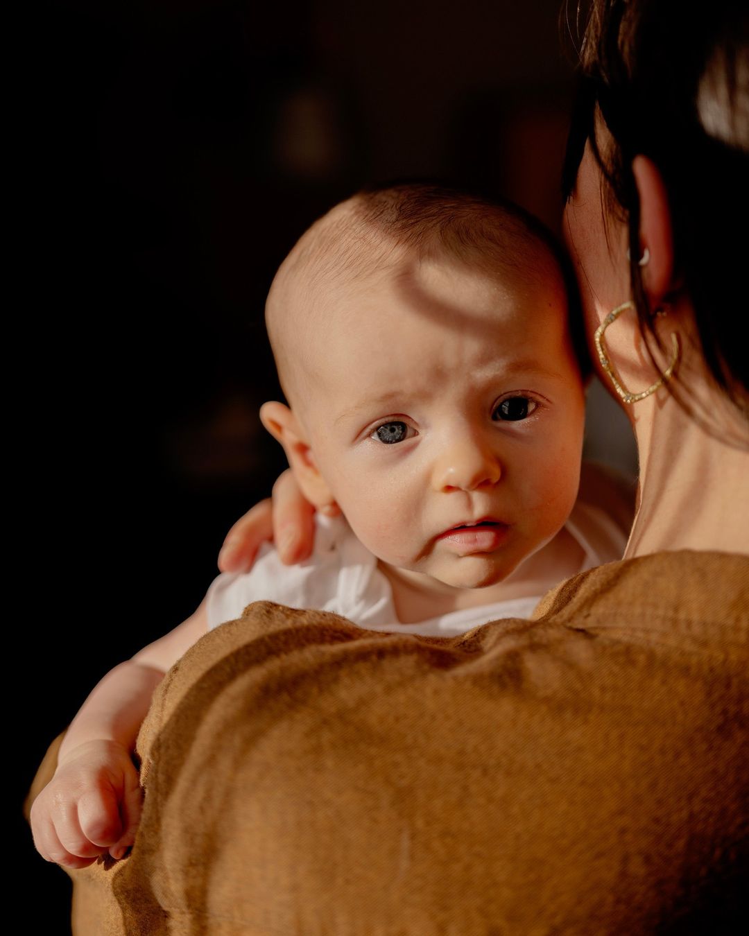 Close-up portrait of a baby being held by a woman in brown clothing, with a dark background highlighting the baby's face.