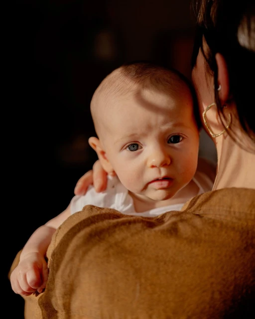 Close-up portrait of a baby being held by a woman in brown clothing, with a dark background highlighting the baby's face.