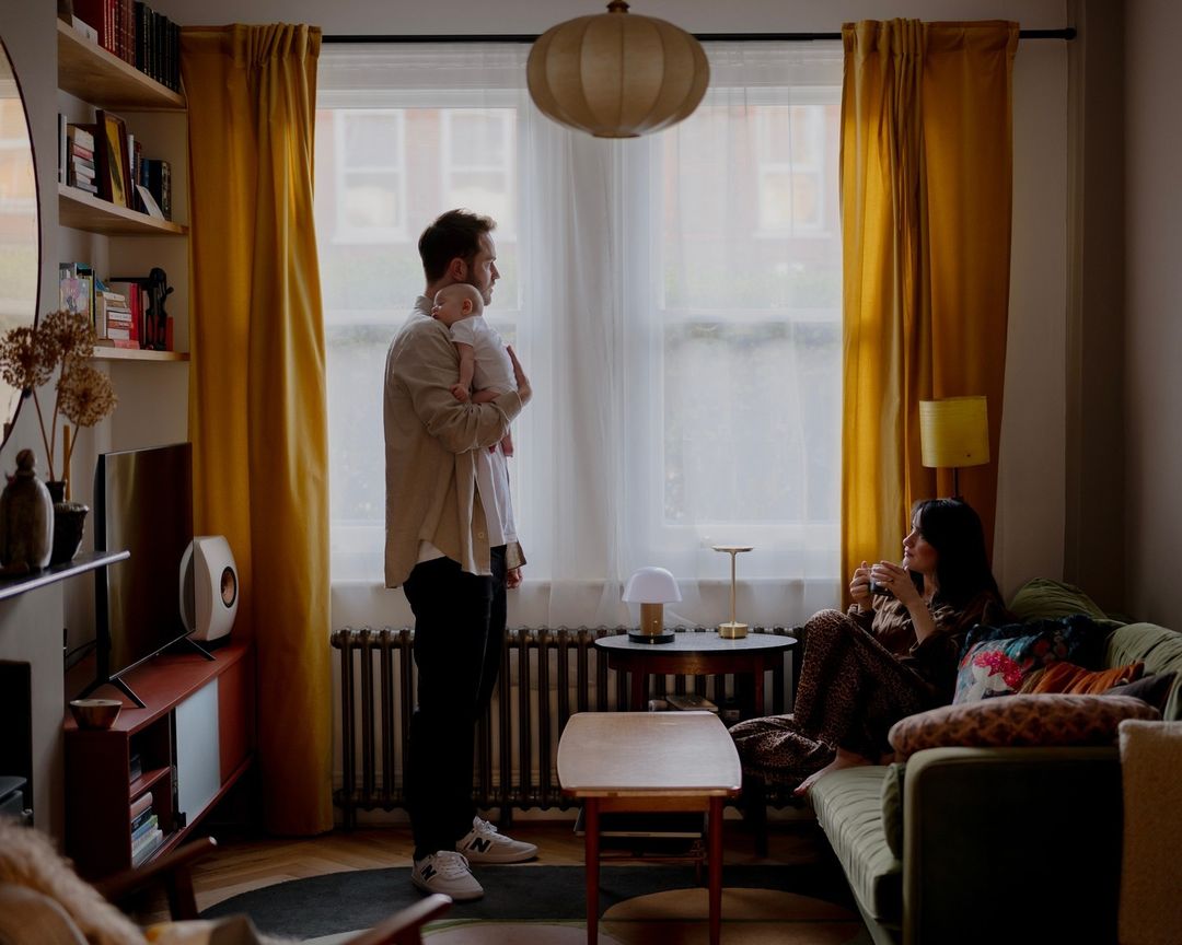 A family of three—a man holding a baby while a woman sits on a couch in a warm, inviting living room with natural light from a window.