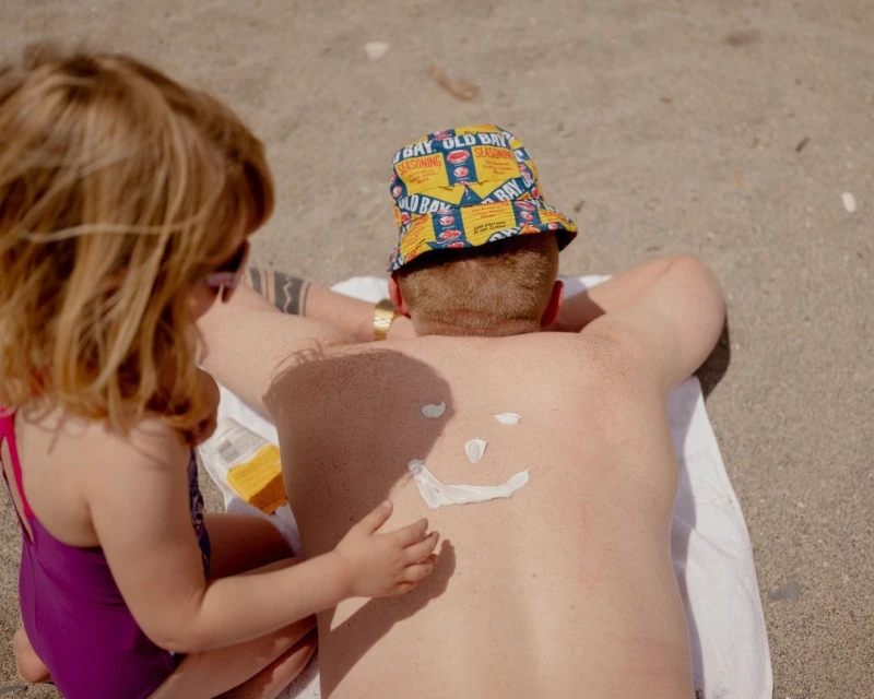 A young child applies sunscreen in a smiley face shape to an adult's back on a sunny beach with the adult wearing a patterned hat.