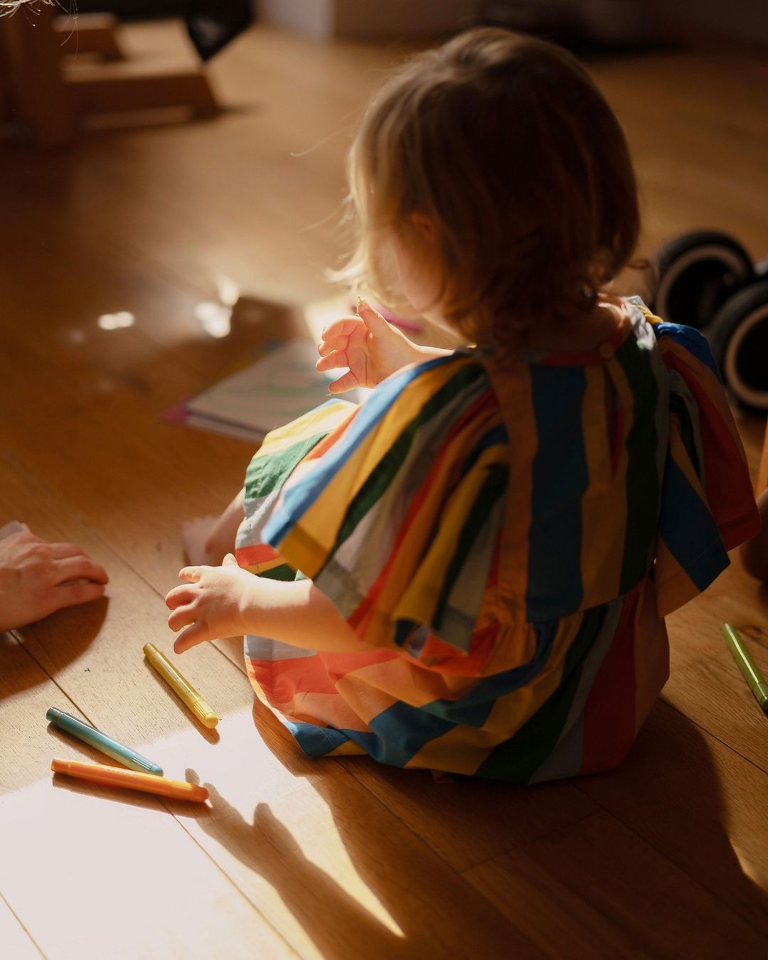 Young child in colorful striped dress sits on a wood floor in dappled sunlight drawing with crayons, back to camera, playful mood.