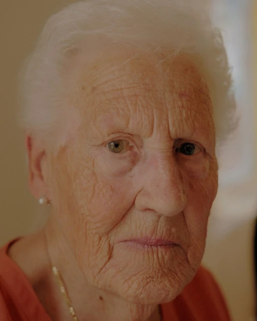 Elderly woman with white hair and neutral expression in a close-up portrait, showcasing detailed skin texture and simple jewelry indoors.