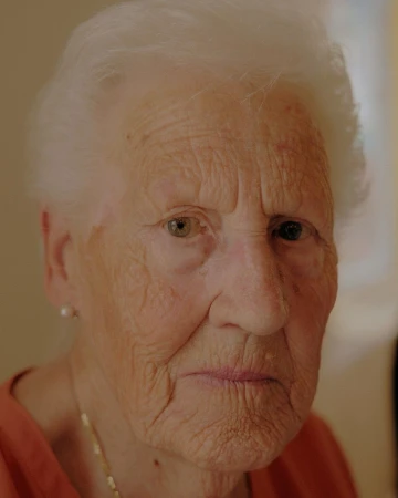 Elderly woman with white hair and neutral expression in a close-up portrait, showcasing detailed skin texture and simple jewelry indoors.