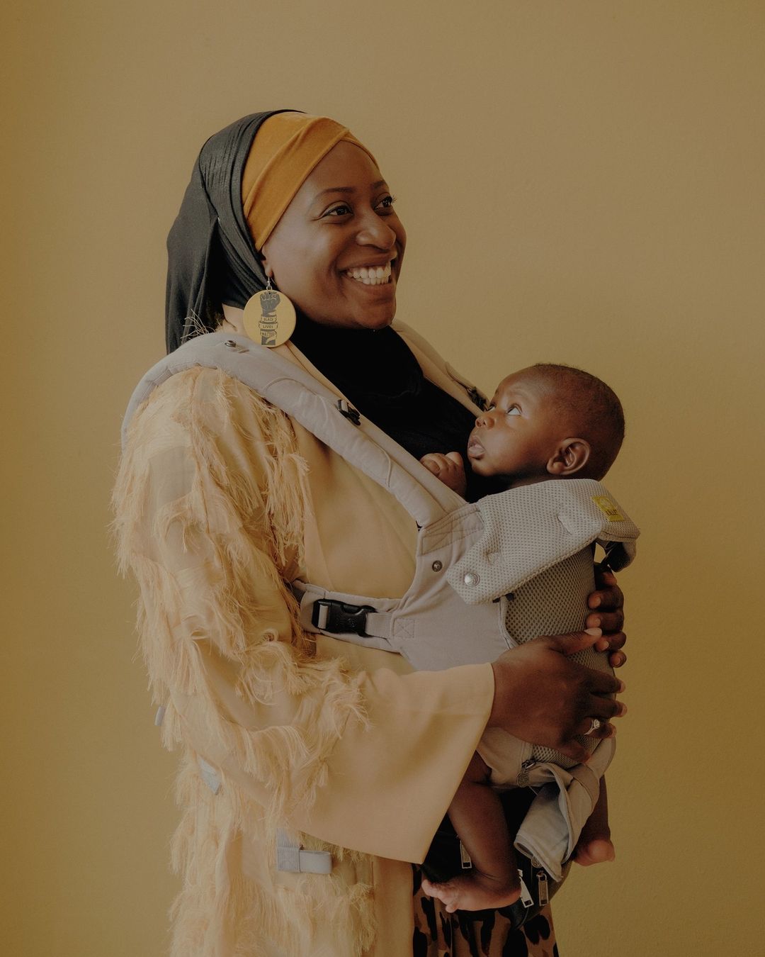 Smiling African-American woman in a fringed coat holding her baby in a carrier against a tan background; studio portrait, family moment.