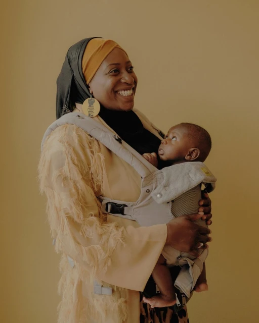 Smiling African-American woman in a fringed coat holding her baby in a carrier against a tan background; studio portrait, family moment.