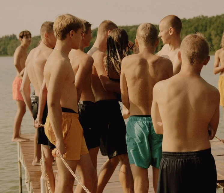 Group of young adult males gathered on a wooden pier near a lake, posing in swim trunks on a sunny day. 