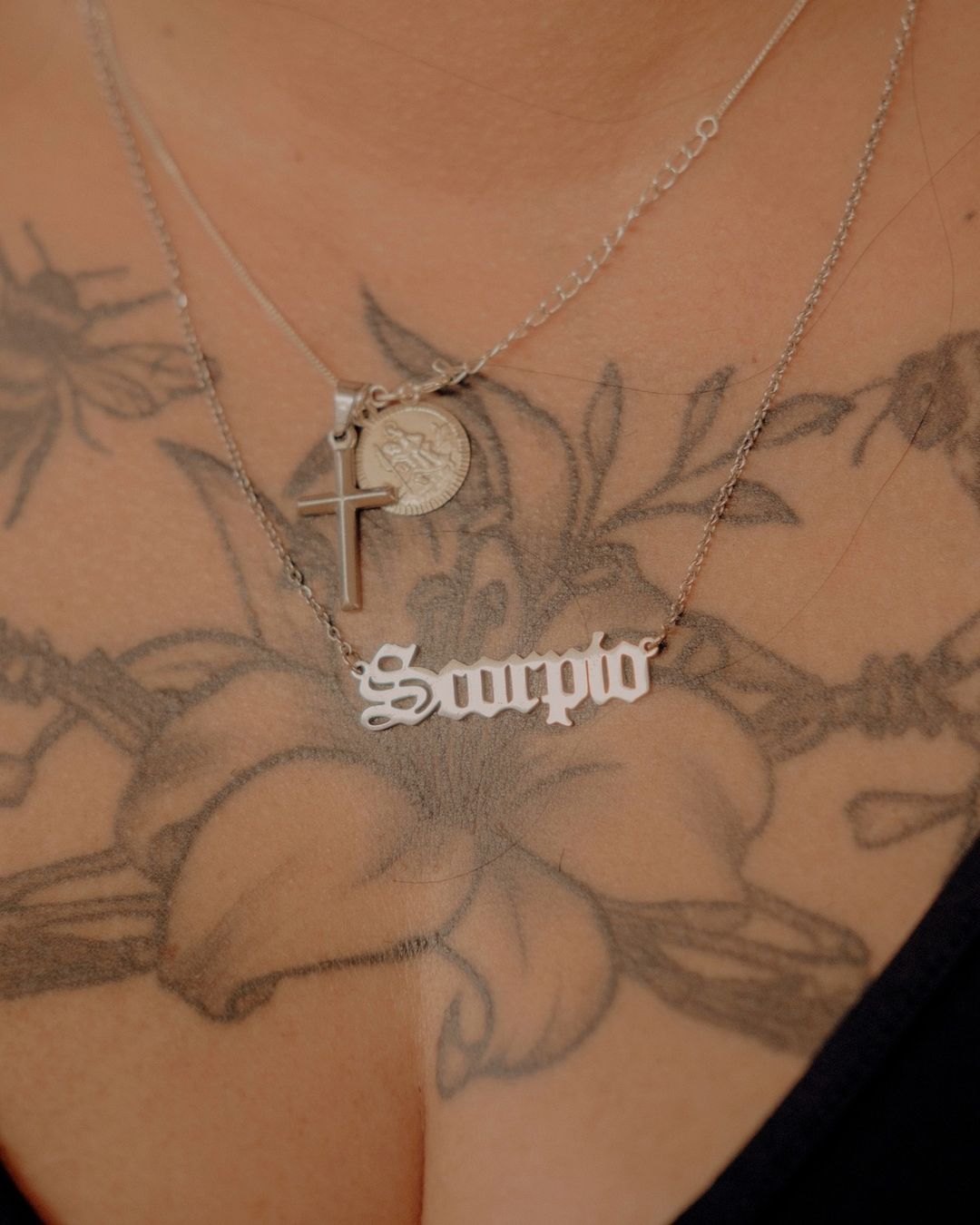 Close-up of a woman's chest with a floral tattoo and two delicate silver necklaces, one featuring a 'Scorpio' pendant.
