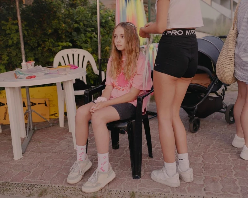 A young girl sits in a chair as a woman braids her hair outdoors with a stroller and another person in the background.