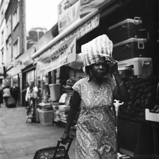Black and white photo of a Black woman walking on a sidewalk with a shopping cart, wearing a head covering on a city street.