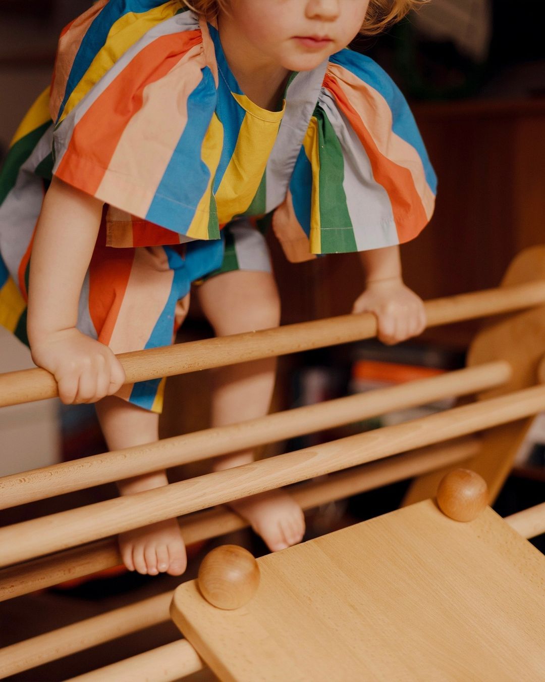 A toddler climbs a wooden play structure indoors, wearing a vibrant striped outfit in blue, yellow, green, orange, and pink.