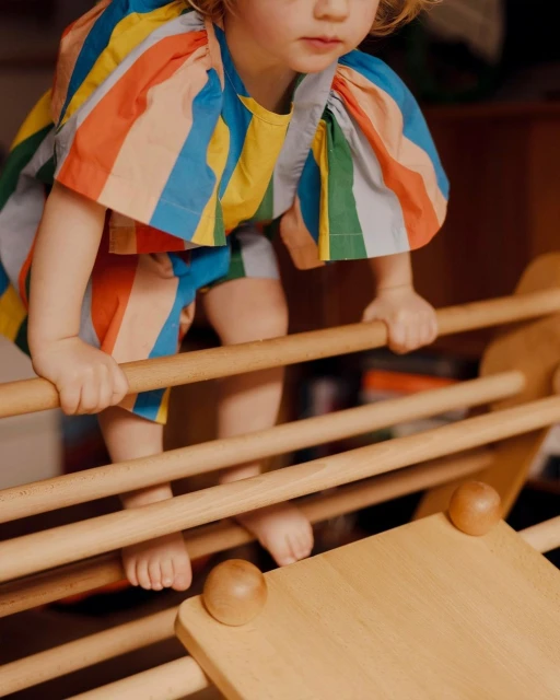 A toddler climbs a wooden play structure indoors, wearing a vibrant striped outfit in blue, yellow, green, orange, and pink.