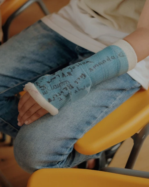 Child sits wearing blue arm cast signed with names on a yellow chair, focusing on healing and community support.