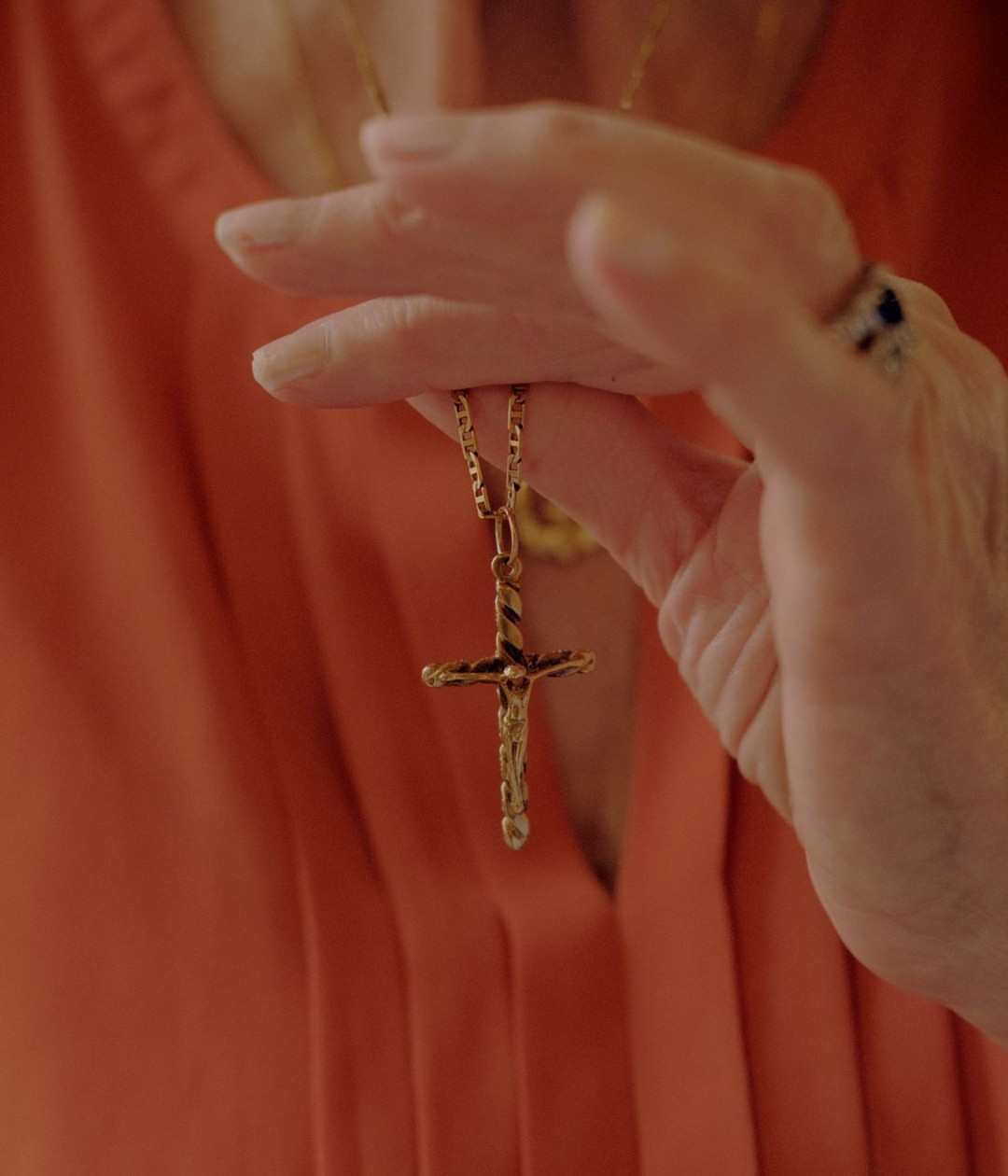 Close-up of an adult holding a gold cross necklace, with a focus on the necklace and hand, set against an orange garment background.