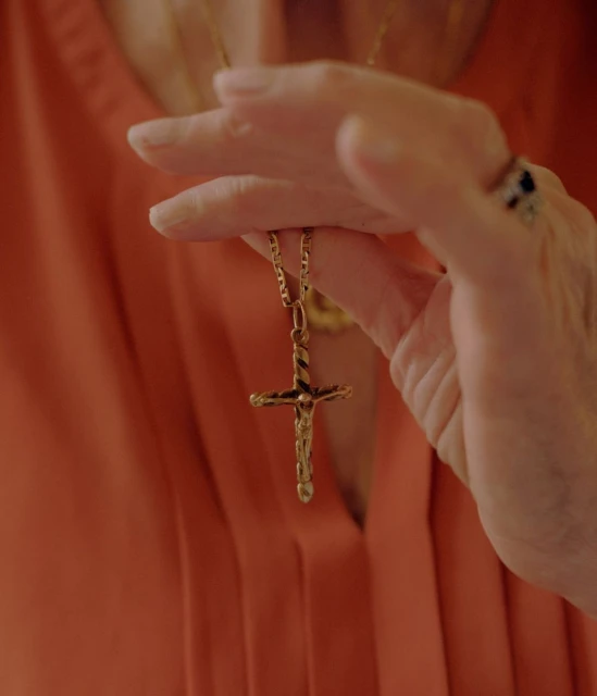 Close-up of an adult holding a gold cross necklace, with a focus on the necklace and hand, set against an orange garment background.