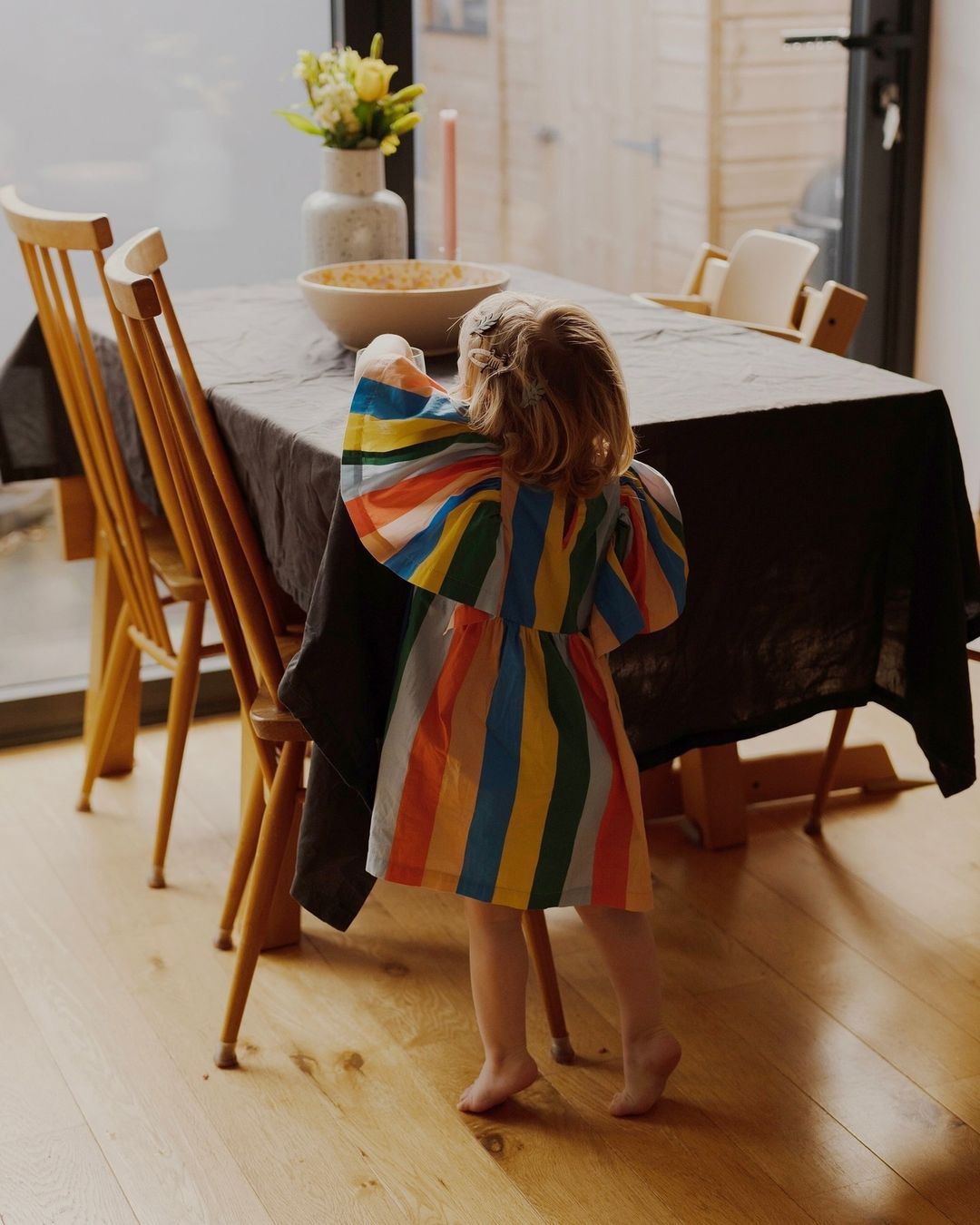 A young child in a colourful striped dress reaches toward a bowl on a dark tablecloth in a dining room captured by Tori Ferenc.