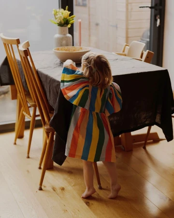 A young child in a colourful striped dress reaches toward a bowl on a dark tablecloth in a dining room captured by Tori Ferenc.