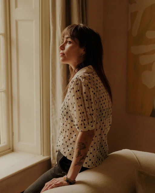 Adult female with long brown hair sitting on a cream couch by a window in a blouse with black dots and bracelets on her arm.