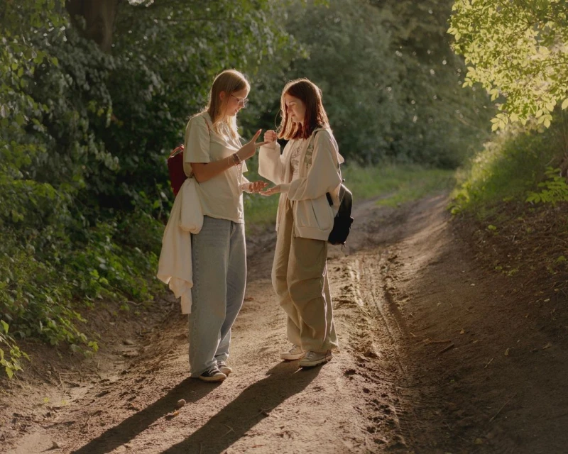 Two young women on a dirt path in a forest, wearing casual clothes and backpacks, engaged in conversation in warm, natural light.