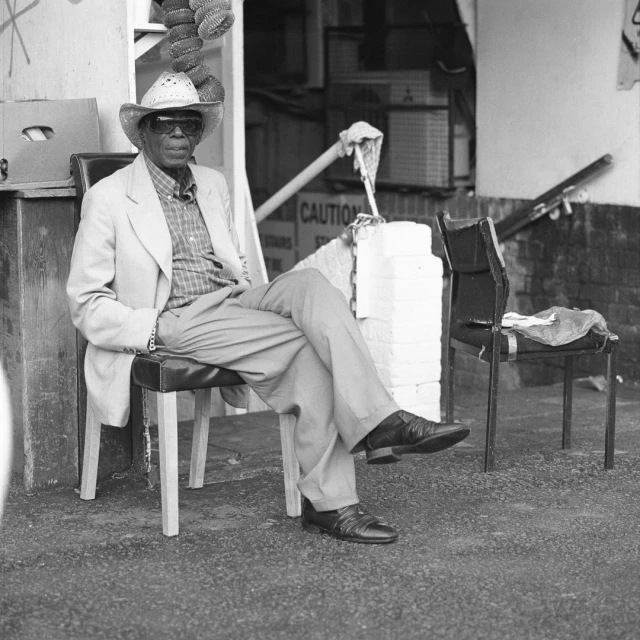 Elderly African-American man in cowboy hat sits outside wearing a suit, with visible caution sign and stacked boxes in background.