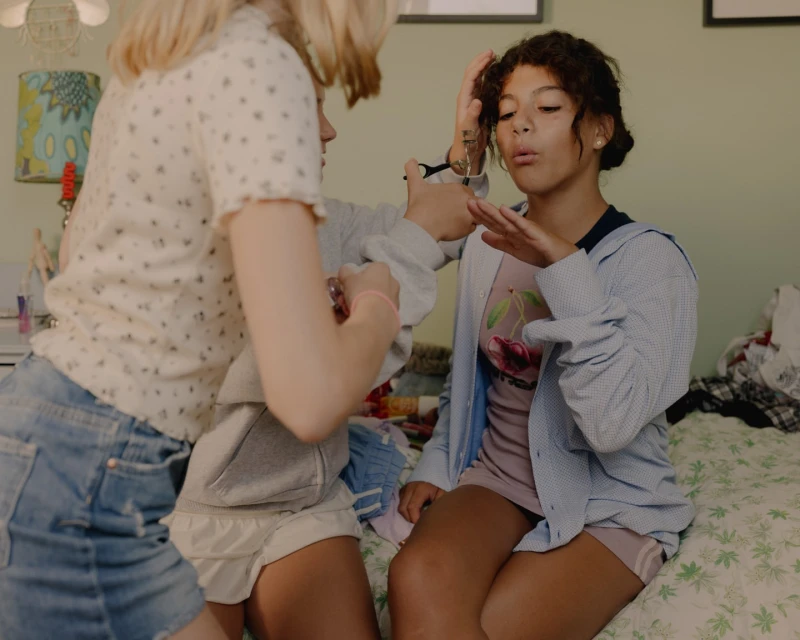 Two young women are indoors on a bed; one appears to be doing the other's makeup or hair in a brightly lit, casual setting.