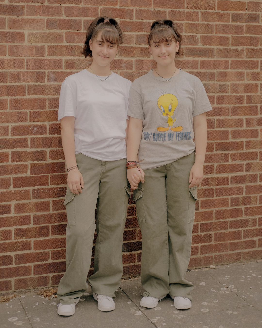 Two young women holding hands pose in front of a brick wall, wearing casual T-shirts, olive cargo pants, and white sneakers, medium shot.